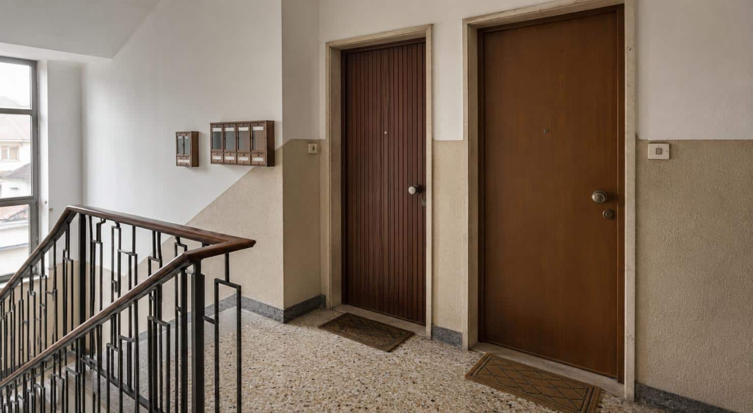 Pianerottolo di un condominio con due porte di appartamento affiancate, zerbini e cassette della posta: le vie di passaggio delle cimici dei letti fra unità confinanti