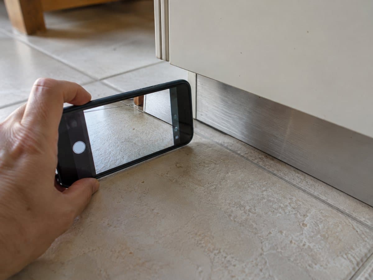 Mano che fotografa con lo smartphone un punto del pavimento chiaro accanto allo zoccolo di un mobile da cucina